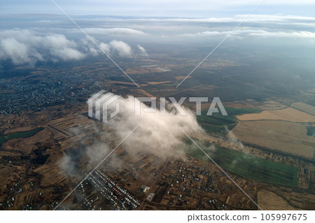 Aerial view from high altitude of distant city covered with puffy cumulus clouds flying by before rainstorm. Airplane point of view of landscape in cloudy weather Aerial view from high altitude of distant city covered with puffy cumulus clouds flying by before rainstorm. Airplane point of view of landscape in cloudy weather 105997675