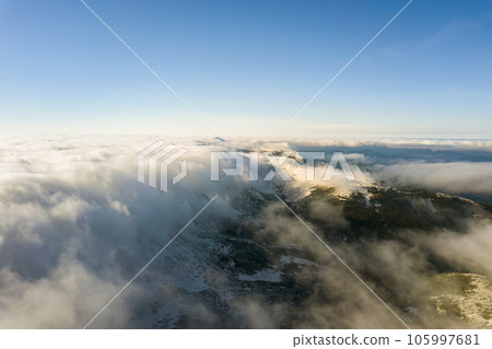 Aerial view from above of white puffy clouds covering snowy mountain tops in bright sunny day. 105997681