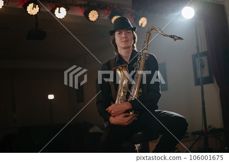 Attractive man with saxophone in his hand sits on chair and looks at camera 106001675