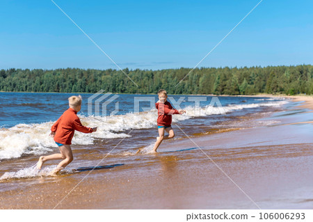 cute caucasian boys wearing red hoodies and blue underpants running from waves in Ladoga lake with enthusiasm. cute caucasian boys wearing red hoodies and blue underpants running from waves in Ladoga lake with enthusiasm. 106006293