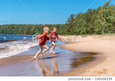 cute caucasian boys wearing red hoodies and blue underpants running from waves in lake with enthusiasm. cute caucasian boys wearing red hoodies and blue underpants running from waves in lake with enthusiasm. 106006294
