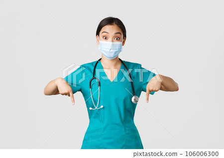 Covid-19, coronavirus disease, healthcare workers concept. Excited and surprised asian female physician, nurse in scrubs and medical mask, pointing fingers down, showing advertisement 106006300