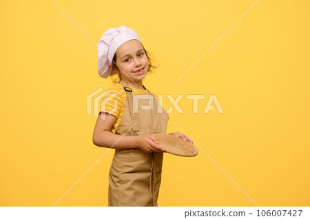 Cute little chef confectioner, cook, baker, pizzaiolo, adorable kid girl holds a wooden board, smiles at camera Cute little chef confectioner, cook, baker, pizzaiolo, adorable kid girl holds a wooden board, smiles at camera 106007427