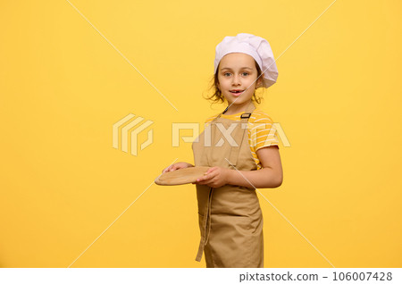 Adorable kid schoolgirl dressed as chef confectioner, holds a wooden board, smiles looking at camera, isolated on yellow 106007428