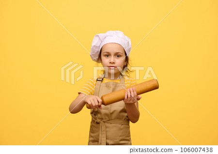 Portrait of a baby girl in chef's uniform, holding a wooden rolling pin, looking at camera, isolated yellow background 106007438