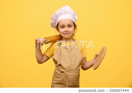 Adorable girl in chef hat and apron, smiles, looking at camera, holding rolling pin and wooden board, isolated on yellow 106007444