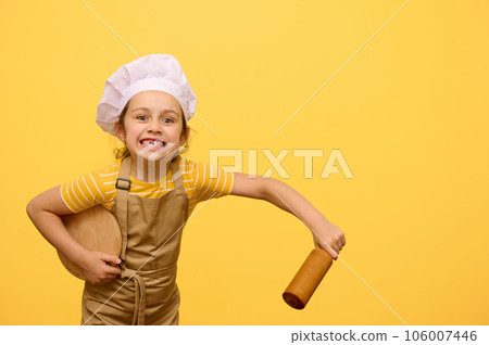 Kid girl in chef hat and apron, makes faces, looking at camera, holding rolling pin and wooden board, isolated on yellow Kid girl in chef hat and apron, makes faces, looking at camera, holding rolling pin and wooden board, isolated on yellow 106007446