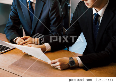 Part cut of the hand of a businessman man in a suit who explains document materials to the person next to him at the meeting table in the office Part cut of the hand of a businessman man in a suit who explains document materials to the person next to him at the meeting table in the office 106008354