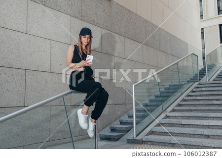 Smiling young woman sitting on stair railing in the city using phone gadget, reading or chatting on smartphone 106012387
