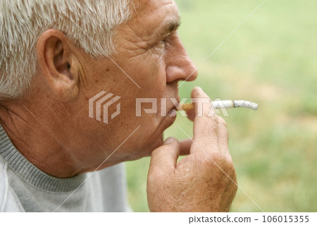 Senior man lighting up a cigarette outdoors against green nature background. Old thoughtful man smoking, looking aside. Close Up Face. Relaxed pensioner enjoy weekend rest outside. Unhealthy habits 106015355