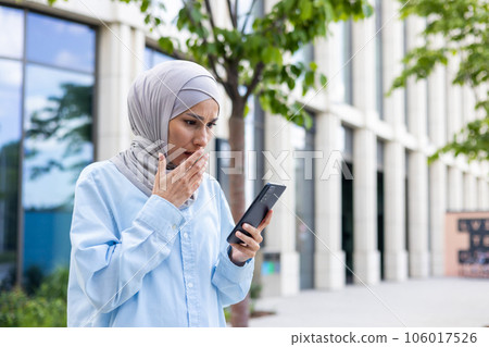 Young business woman in hijab walking in the city, Arab woman received online notification with bad news, holding phone in hands reading upset and depressed sad. 106017526
