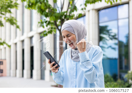 Joyful woman in hijab walking in the city, received notification of online win and good news on phone, business woman outside office building, holding hand up triumph winner gesture. 106017527