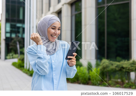 Joyful woman in hijab walking in the city, received notification of online win and good news on phone, business woman outside office building, holding hand up triumph winner gesture. Joyful woman in hijab walking in the city, received notification of online win and good news on phone, business woman outside office building, holding hand up triumph winner gesture. 106017529