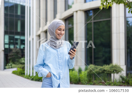 A young beautiful business woman in a hijab walks through the city, a Muslim woman holds a phone in her hands, uses an application on a smartphone, smiles, browses the Internet. A young beautiful business woman in a hijab walks through the city, a Muslim woman holds a phone in her hands, uses an application on a smartphone, smiles, browses the Internet. 106017530