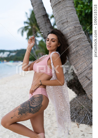 Stylish young woman with voluminous hair in a bikini and lace pareo posing on a tropical beach, slim, tanned, athletic, sunglasses 106018318