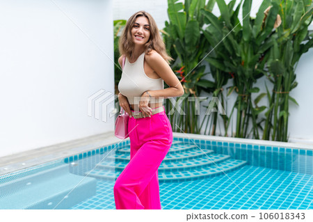 Stylish fit fashion women in bright pink wide leg pants holding bag trendy mint sunglasses posing at luxury tropical villa by pool outdoor natural day light 106018343