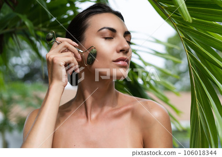 Beauty portrait, soft skin. Female model with natural make-up and healthy skin behind a green leafy plant. Portrait of a beautiful girl with nude nails, bare shoulders holding roller massager outdoor Beauty portrait, soft skin. Female model with natural make-up and healthy skin behind a green leafy plant. Portrait of a beautiful girl with nude nails, bare shoulders holding roller massager outdoor 106018344
