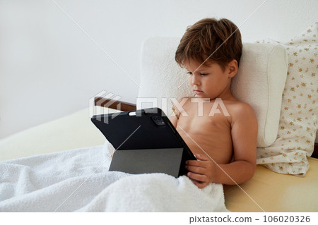 A sick child looks at a tablet while measuring temperature with a mercury thermometer. A sick child looks at a tablet while measuring temperature with a mercury thermometer. 106020326