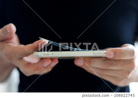 Process unrecognisable repairman is removing broken and old tempered glass screen protector from smartphone, close up view. Services for gluing and replacement of damaged protective glass. Process unrecognisable repairman is removing broken and old tempered glass screen protector from smartphone, close up view. Services for gluing and replacement of damaged protective glass. 106022745