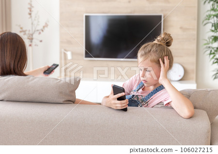 Girl sitting on couch and watching something interesting in mobile phone while mother watching tv Girl sitting on couch and watching something interesting in mobile phone while mother watching tv 106027215