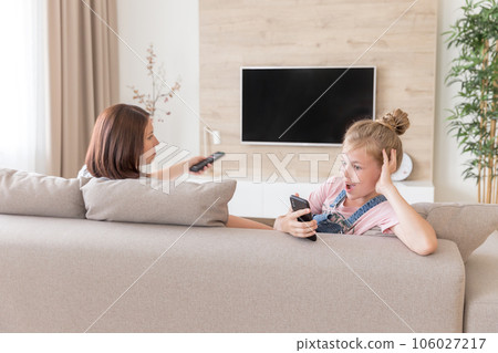 Girl sitting on couch and watching something interesting in mobile phone while mother watching tv. surprise concept Girl sitting on couch and watching something interesting in mobile phone while mother watching tv. surprise concept 106027217