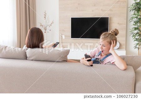 Girl sitting on couch and watching something interesting in mobile phone while mother watching tv. surprise concept Girl sitting on couch and watching something interesting in mobile phone while mother watching tv. surprise concept 106027230