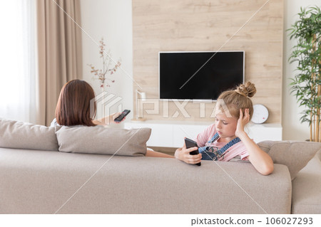 Girl sitting on couch and watching something interesting in mobile phone while mother watching tv 106027293