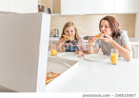 Mother and daughter sitting in the kitchen, eating pizza and having fun. Focus on daughter Mother and daughter sitting in the kitchen, eating pizza and having fun. Focus on daughter 106028067