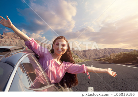 Happy woman smiling and holding hands outside open window car against road and mountain background.  106039196