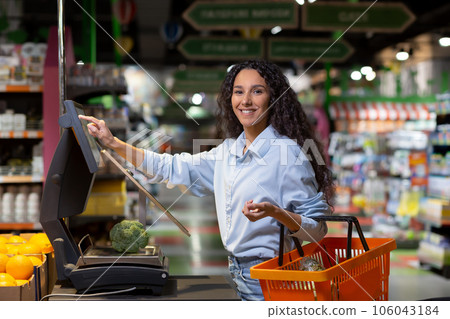 Portrait of satisfied female shopper in supermarket, Hispanic woman weighing fruit in store, smiling and looking at camera using self-service scales. 106043184