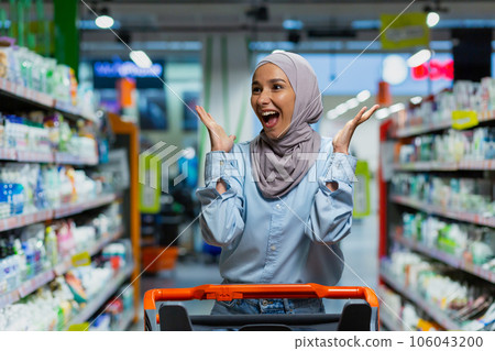 Portrait of successful satisfied woman shopper in supermarket, cheerful muslim woman in hijab with shopping trolley among shelves with goods looking at various shops. 106043200