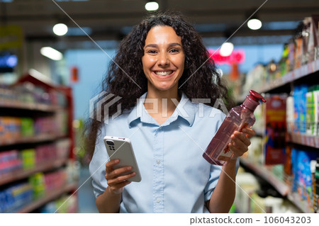 Portrait of a female buyer in a supermarket, a Hispanic woman chooses shampoo for hair and shower gel, reads reviews on the Internet, smiles and uses an application on her phone. Portrait of a female buyer in a supermarket, a Hispanic woman chooses shampoo for hair and shower gel, reads reviews on the Internet, smiles and uses an application on her phone. 106043203
