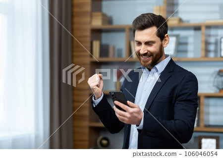 A mature businessman in the office received an online notification of a victory win, the winner holds his hand up a gesture of triumph, a man holds a phone in his hands, uses an application. 106046054