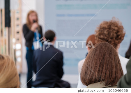 Back view of group of people gathering together in conference hall and listening to presentation of female speaker with microphone Back view of group of people gathering together in conference hall and listening to presentation of female speaker with microphone 106046666