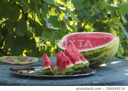Ripe red watermelon and slices on the table in front of green leaves; sunlight, Ripe red watermelon and slices on the table in front of green leaves; sunlight, 106049114