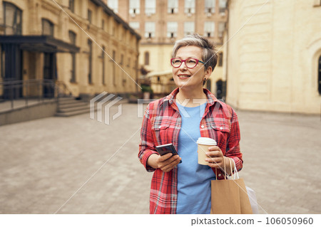 Smiling mature woman walking with her shopping bags, drinking coffee and use phone in city street  106050960