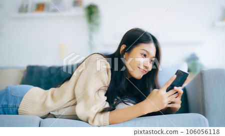Happy young asian woman relax on comfortable couch at home texting messaging on smartphone 106056118