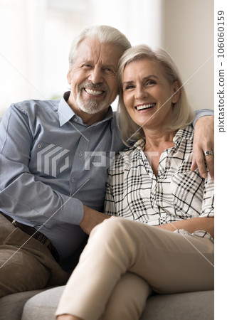 Cheerful positive senior retired husband and wife hugging on sofa 106060159