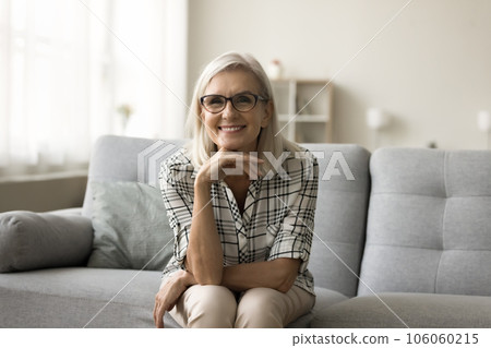 Happy blonde mature woman in stylish eyeglasses sitting on couch Happy blonde mature woman in stylish eyeglasses sitting on couch 106060215