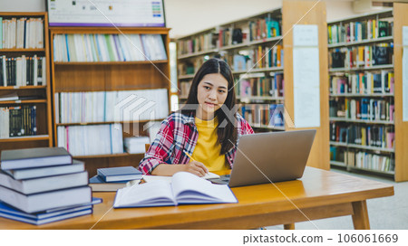 Female college student working on laptop and searching for books to study, make report, find useful information in college room. Concept of reading, learning and intelligence. 106061669