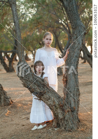 Two sisters, kid and teenage girl in white clothes standing near tree in the park Two sisters, kid and teenage girl in white clothes standing near tree in the park 106063817