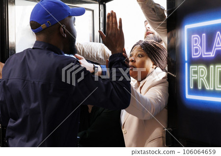 Rude aggressive shoppers try to enter into shopping mall and get best Black Friday deals first. Store security officer holding back angry crazy crowd of diverse people breaking into store during sales 106064659