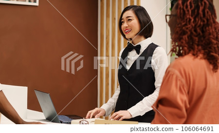 Happy friendly asian receptionist helping guests during check in process, imputing their personal information on laptop. Tourists booking in their stay in luxury hotel, assisted by hotel staff 106064691