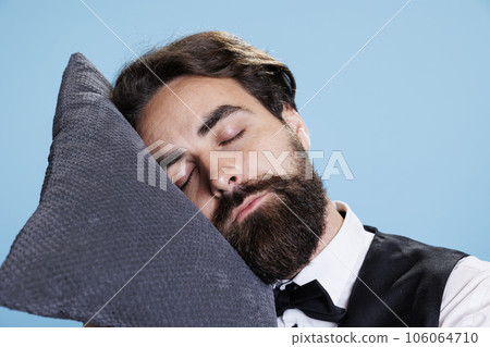 Classy bellboy falling asleep on pillow while he stands in studio, feeling extremely worn out and exhausted. Professional employee doorman taking a quick nap on camera, expressing burnout. Classy bellboy falling asleep on pillow while he stands in studio, feeling extremely worn out and exhausted. Professional employee doorman taking a quick nap on camera, expressing burnout. 106064710