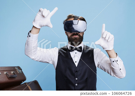 Modern doorkeeper using vr headset against blue background, enjoying 3d interactive vision experience. Professional bellboy hotel worker having fun with virtual reality glasses. Modern doorkeeper using vr headset against blue background, enjoying 3d interactive vision experience. Professional bellboy hotel worker having fun with virtual reality glasses. 106064726
