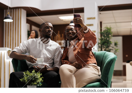 Happy guests taking photos in hotel lobby, relaxing in lounge area before accommodation. Man and woman having fun with picutres on smartphone, doing check in at luxury resort. Happy guests taking photos in hotel lobby, relaxing in lounge area before accommodation. Man and woman having fun with picutres on smartphone, doing check in at luxury resort. 106064739