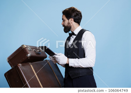 Hotel concierge counting trolley bags and checking tablet with list of reservations, studio shot with blue background. Young professional doorman with gloves taking care of luggage. Hotel concierge counting trolley bags and checking tablet with list of reservations, studio shot with blue background. Young professional doorman with gloves taking care of luggage. 106064749