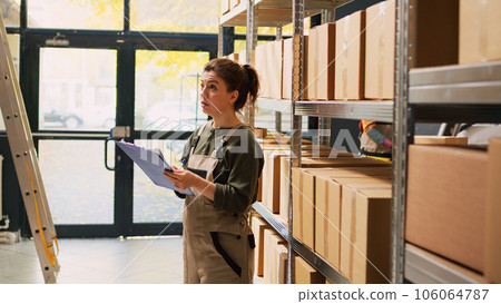 Depot worker checking stock inventory, working on logistics inspection to ship products orders from storage room. Woman managing boxes of cargo goods with list for quality control. 106064787