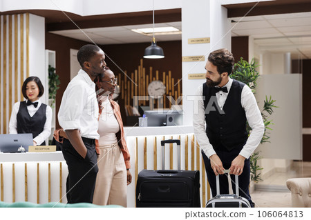 Hotel steward carrying bags at resort, offering to help guests with their luggage at reception desk in lobby. Bellboy providing excellent luxury service to tourists, trolley bags. 106064813
