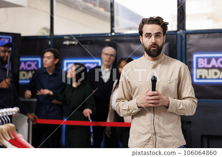 Young man journalist making news about Black Friday biggest shopping event, talking over microphone against crowd of shoppers. TV reporter live broadcasting from shopping mall during sales season Young man journalist making news about Black Friday biggest shopping event, talking over microphone against crowd of shoppers. TV reporter live broadcasting from shopping mall during sales season 106064860